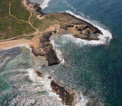 An aerial view of the Sinis peninsula coastline showing an encurved bay with sandy beaches and rocky headlands jutting into deep blue water. White foam from waves breaks against the rocky jagged coast, while green vegetation covers the undulating coastal terrain. Winding dirt paths are visible crossing the headland above the shore.