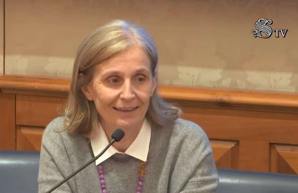 Our scientific coordinator Annalisa Falace seated at a press conference table in the Italian Senate chamber. She is speaking into a microphone, wearing a grey sweater over a white collared shirt with a colorful beaded necklace. The formal Senate setting features wood-paneled walls and blue upholstered seating. The Senate TV logo is visible in the upper right corner.