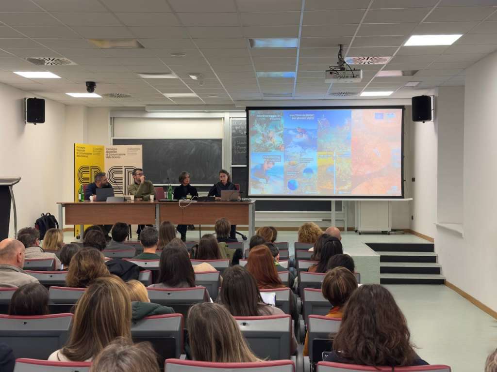 The scene shows a modern university lecture hall with tiered seating filled with an engaged audience of approximately 40-50 attendees. At the front of the room, three panelists are seated behind a wooden presentation table with laptops and water bottles. Behind them, a large projection screen displays some of the contents realized for the REEForest social media pages.