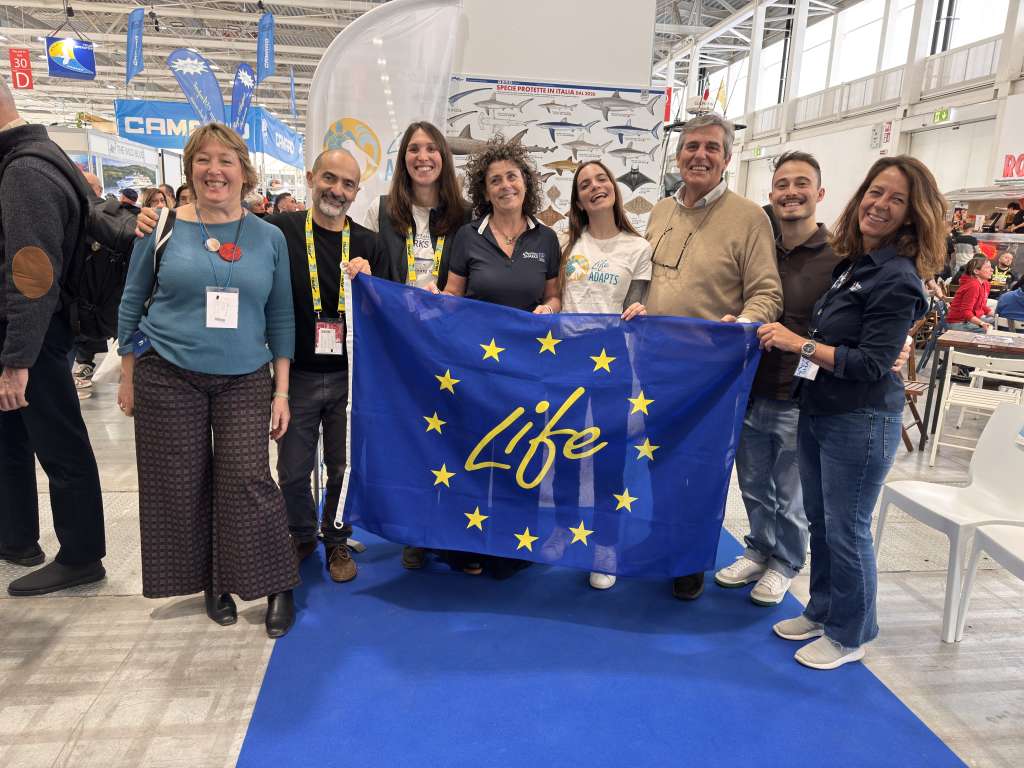 A group of eight smiling adults poses together at the EUDI exhibition hall, proudly holding a large blue European Union flag with the word "Life" written in gold script across the stars. Some participants wear lanyards and project badges. A display panel about shark species protected in Italy is visible in the background.