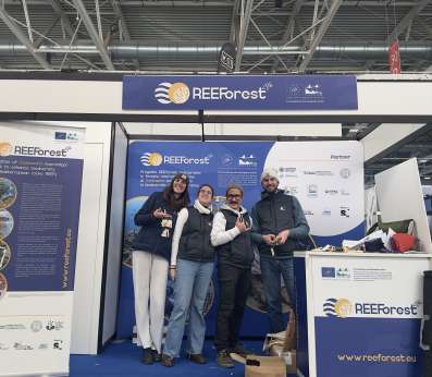 Four members of the LIFE REEForest project team stand side by side at their exhibition booth, smiling at the camera, shortly before the opening of the fair. Behind them, the REEForest branded backdrop explains the project's goal of restoring Cystoseira macroalgal forests to enhance Mediterranean biodiversity. The booth banner with the project logo hangs overhead.
