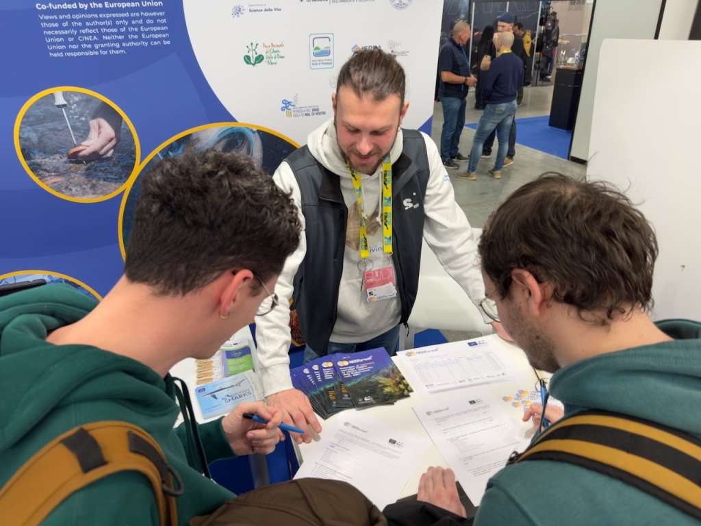 A diver and researcher from the LIFE REEForest project stands behind an exhibition table covered in informational brochures and questionnaires, leaning forward to assist two young male visitors who are filling out forms. The booth backdrop features the EU co-funding logo and circular photos of marine research activities.
