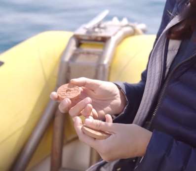 In the foreground, a woman's hands holding two clay disks used to grow new algae of the Cystoseira sensu lato group. She is standing on a yellow boat and the sea can be seen in the background.
