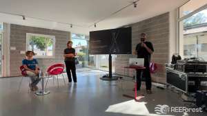 A bright, modern indoor venue with large windows in the library of Staranzano hosts a small presentation. Francesco Scarel dressed in black, organizer of the Festival dell'Acqua, holds a microphone and speaks beside a laptop on a red stand, next to a large screen displaying a dark slide credited to artist Miha Godec, with the subtitle "Art, science, and ecology in dialogue asking what coexistence might look like in the future." A man in black stands to his left with arms crossed, listening. Elena Toti, wearing a blue REEForest-branded t-shirt sits on a red chair to the left, also preparing for the talk. Audio and technical equipment cases are visible to the right. Sunlight streams in through the floor-to-ceiling windows.