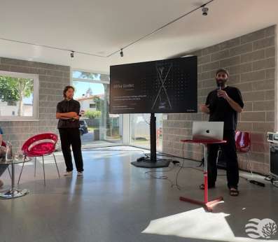 A bright, modern indoor venue with large windows in the library of Staranzano hosts a small presentation. Francesco Scarel dressed in black, organizer of the Festival dell'Acqua, holds a microphone and speaks beside a laptop on a red stand, next to a large screen displaying a dark slide credited to artist Miha Godec, with the subtitle "Art, science, and ecology in dialogue asking what coexistence might look like in the future." A man in black stands to his left with arms crossed, listening. Elena Toti, wearing a blue REEForest-branded t-shirt sits on a red chair to the left, also preparing for the talk. Audio and technical equipment cases are visible to the right. Sunlight streams in through the floor-to-ceiling windows.