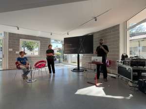 A bright, modern indoor venue with large windows in the library of Staranzano hosts a small presentation. Francesco Scarel dressed in black, organizer of the Festival dell'Acqua, holds a microphone and speaks beside a laptop on a red stand, next to a large screen displaying a dark slide credited to artist Miha Godec, with the subtitle "Art, science, and ecology in dialogue asking what coexistence might look like in the future." A man in black stands to his left with arms crossed, listening. Elena Toti, wearing a blue REEForest-branded t-shirt sits on a red chair to the left, also preparing for the talk. Audio and technical equipment cases are visible to the right. Sunlight streams in through the floor-to-ceiling windows.