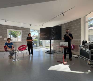 A bright, modern indoor venue with large windows in the library of Staranzano hosts a small presentation. Francesco Scarel dressed in black, organizer of the Festival dell'Acqua, holds a microphone and speaks beside a laptop on a red stand, next to a large screen displaying a dark slide credited to artist Miha Godec, with the subtitle "Art, science, and ecology in dialogue asking what coexistence might look like in the future." A man in black stands to his left with arms crossed, listening. Elena Toti, wearing a blue REEForest-branded t-shirt sits on a red chair to the left, also preparing for the talk. Audio and technical equipment cases are visible to the right. Sunlight streams in through the floor-to-ceiling windows.