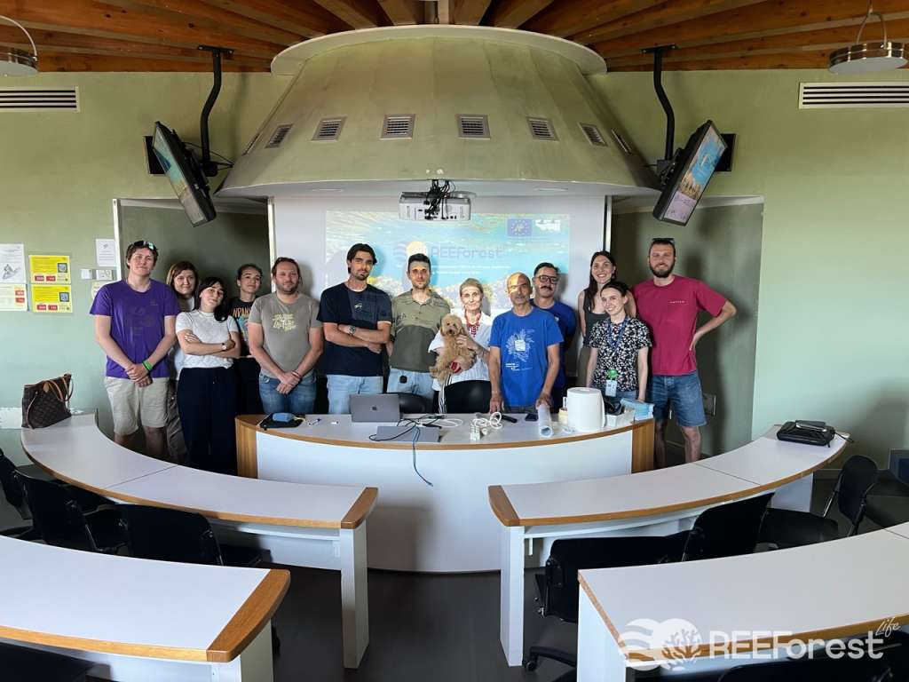 A group of students from the Master's degree in Marine Sciences at the University of Trieste and researchers from the REEForest team pose together at the front of a small circular lecture room, standing behind a curved seminar desk. The REEForest Life project logo and an underwater habitat image are projected on the screen behind them. Professor Annalisa Falace, at the center of the group, holds a small curly-haired dog. The room has a distinctive round ceiling structure with wooden beams and multiple wall-mounted screens.