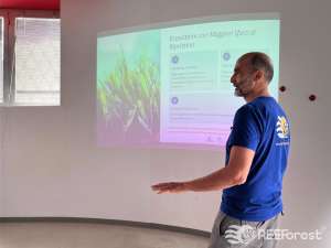 Saul Ciriaco from the Shoreline team, wearing a blue REEForest-branded t-shirt, stands sideways in front of a projected slide titled Ecosistemi con Maggiori Sforzi di Ripristino (Ecosystems with the Greatest Restoration Efforts). The slide lists three marine ecosystem types — Posidonia oceanica meadows, reef corals, and Cystoseira sensu lato forests — with statistics on the proportion of restoration efforts dedicated to each. An underwater image of green seagrass is projected on the left side of the screen.
