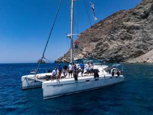 Nine REEForest team members stands together on the bow of a white catamaran named Athos, moored in a sheltered bay with deep blue sea and a steep, arid rocky hillside behind them. The team is dressed casually in white and blue t-shirts, some wearing sunglasses and hats. Flags fly from the mast above. The image conveys a successful field campaign day, with the research vessel serving as the base of operations for Cystoseira s.l. restoration activities in the open sea.