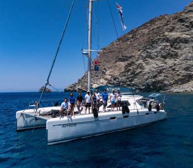 Nine REEForest team members stands together on the bow of a white catamaran named Athos, moored in a sheltered bay with deep blue sea and a steep, arid rocky hillside behind them. The team is dressed casually in white and blue t-shirts, some wearing sunglasses and hats. Flags fly from the mast above. The image conveys a successful field campaign day, with the research vessel serving as the base of operations for Cystoseira s.l. restoration activities in the open sea.