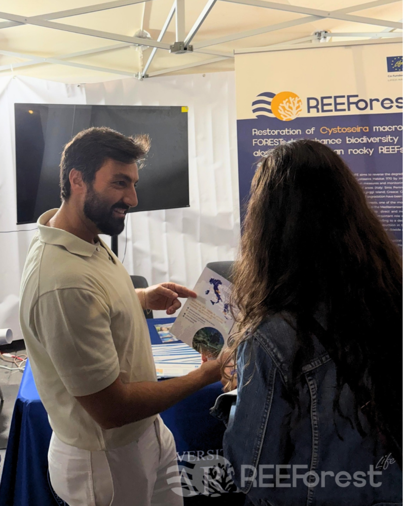 In the forefront, our researcher Dr. Francesco Rendina is showing one of our flyers to a woman with her back turned to the camera. He is smiling while talking about the project. In the background, a roll-up of the REEForest project can be seen on the left side, while a turned off TV screen is visible on the right side.